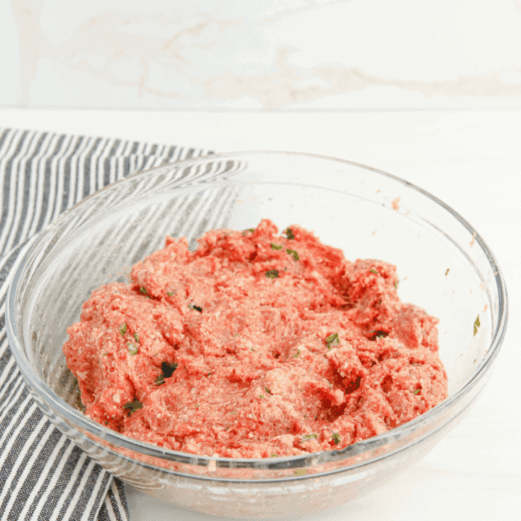 Hands forming evenly sized meatballs from seasoned meat mixture on a tray.