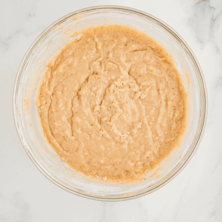 Batter mixture with mashed bananas, eggs, sugars, milk, and vanilla being whisked together beside a bowl of dry ingredients—flour, cardamom, and cinnamon.