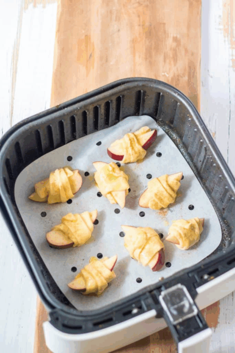Air fryer basket lined with parchment paper, rolled apple crescent rolls spaced evenly.