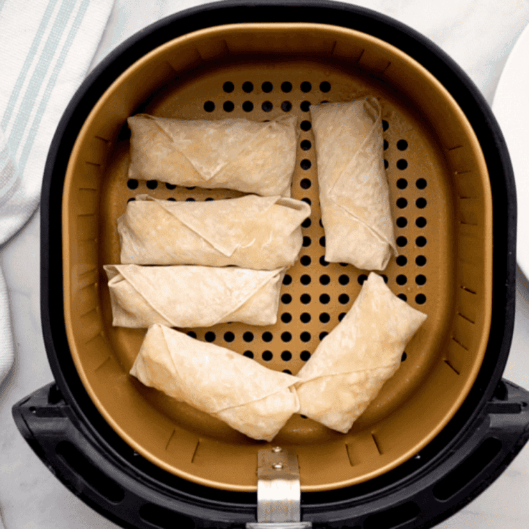 Breakfast egg rolls being placed in a single layer into the air fryer basket after being sprayed with cooking oil for even browning and crisping