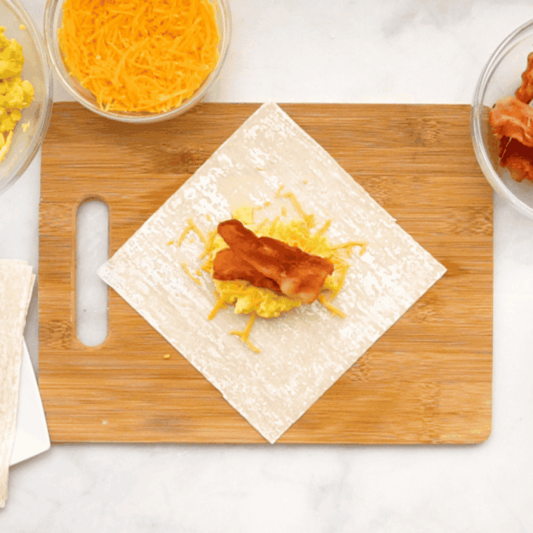 Hands folding an egg roll wrapper over a spoonful of breakfast filling, demonstrating how to tightly roll and seal the sides and corner.