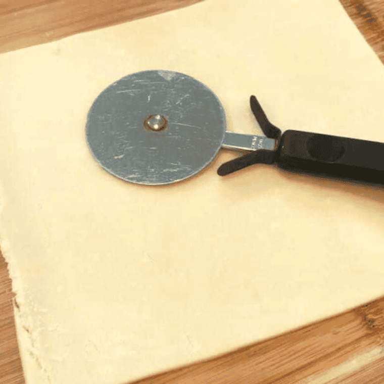 Pie dough laid flat on a surface, being trimmed and cut into a square with a pizza cutter.