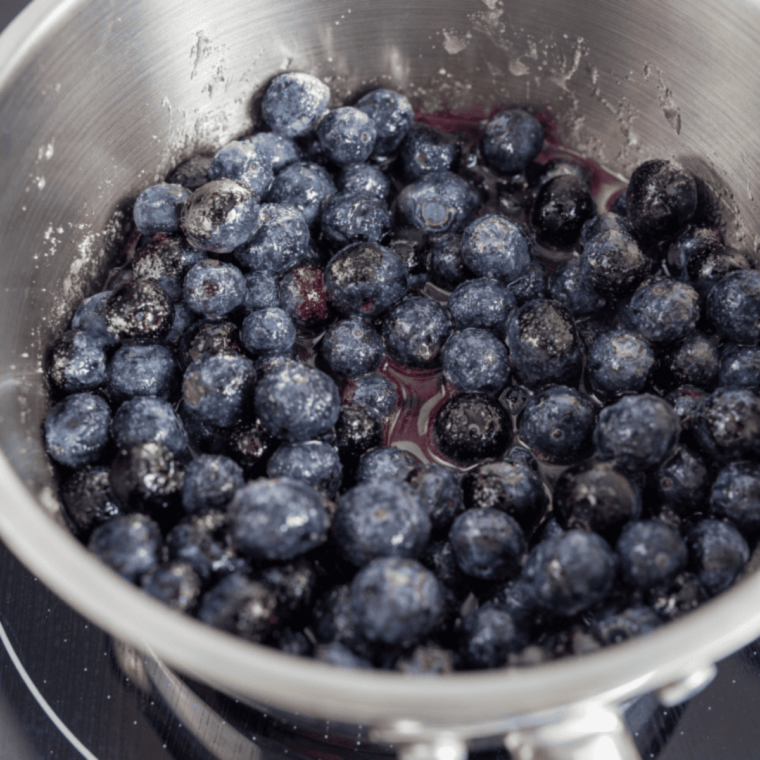 Blueberries, sugar, and lemon zest cooking in a saucepan with cornstarch slurry being added to thicken the mixture.
