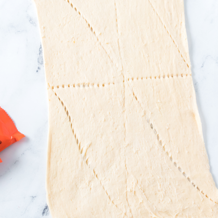 Crescent roll dough is unrolled on a lightly floured surface and separated into triangles or squares. Working with cool dough helps it stay less sticky and easier to wrap around the candy.