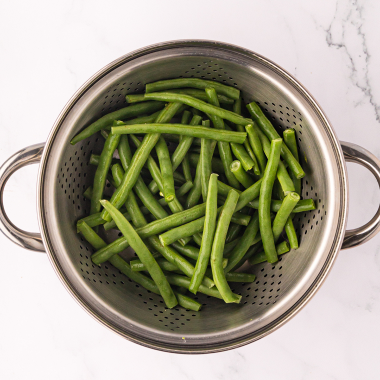 Fresh green beans rinsed in a colander, then trimmed and cut into 2-inch pieces on a cutting board