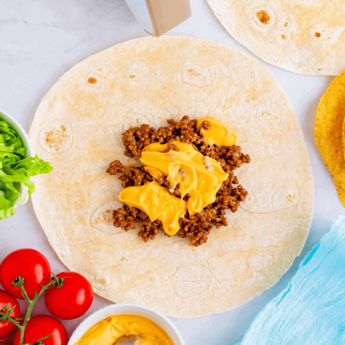 Large tortilla on a work surface topped with seasoned ground beef and nacho cheese, starting layers for air fryer crunchwrap.