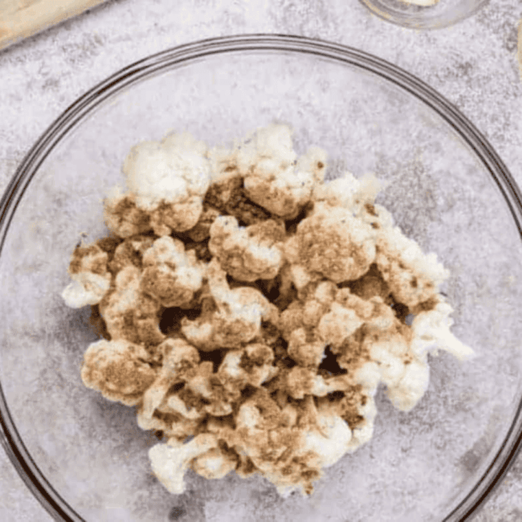 Seasoning fresh cauliflower florets in a bowl with white pepper, garlic powder, cumin, paprika, dry mustard, and black pepper for flavorful Buffalo cauliflower bites.