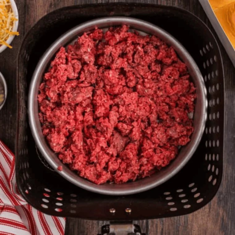 Ground beef being stirred with a spatula inside an air fryer basket halfway through the 10-minute cooking cycle to ensure even browning.