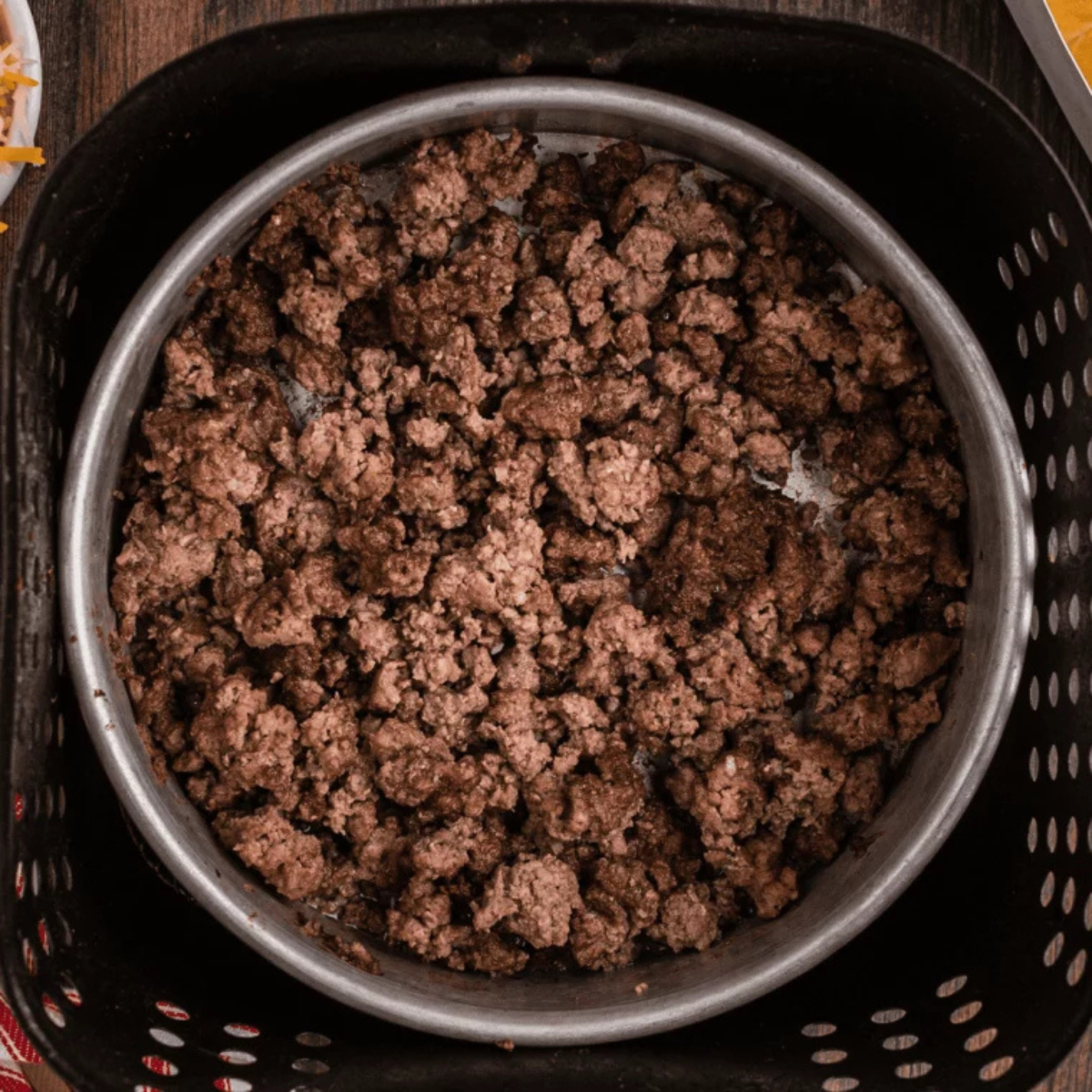 Inserting a meat thermometer into fully browned ground beef in an air fryer basket to ensure it has reached a safe internal temperature of 160&deg;F.