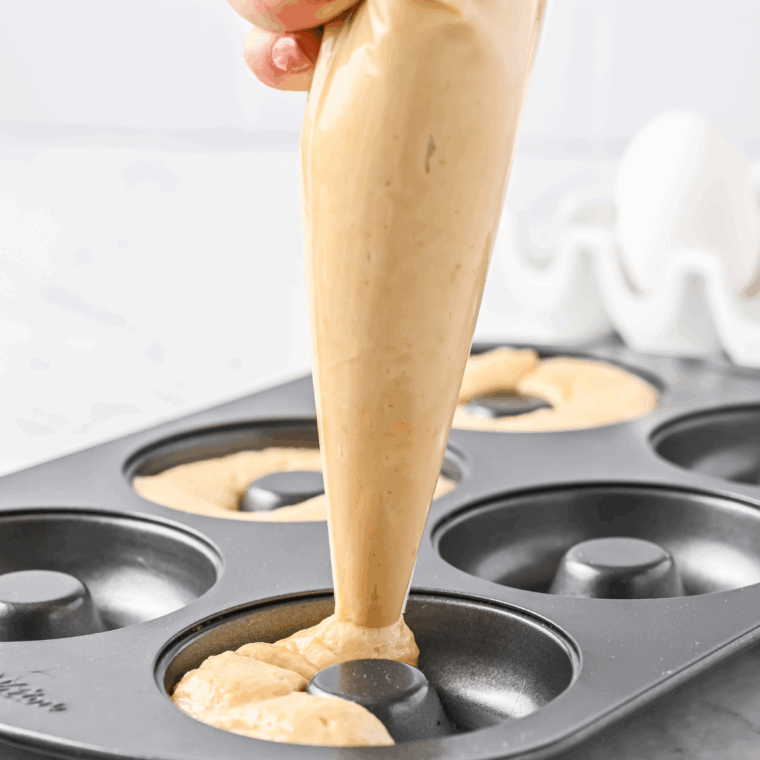 Piping donut batter evenly into six prepared donut pan cavities using a piping bag or plastic bag.