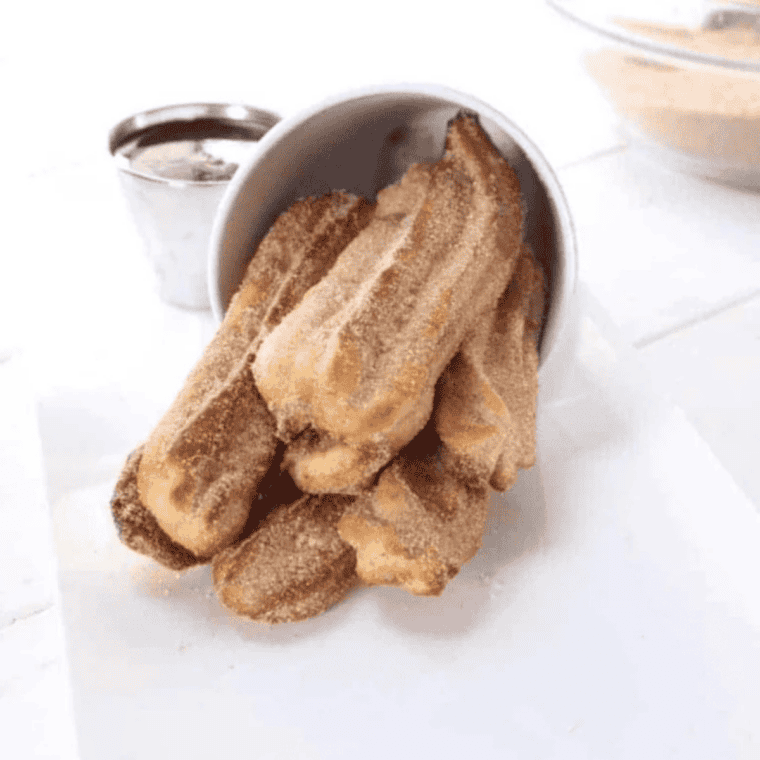 Golden air fryer churros coated in cinnamon sugar stacked on parchment paper with a small bowl of chocolate dipping sauce