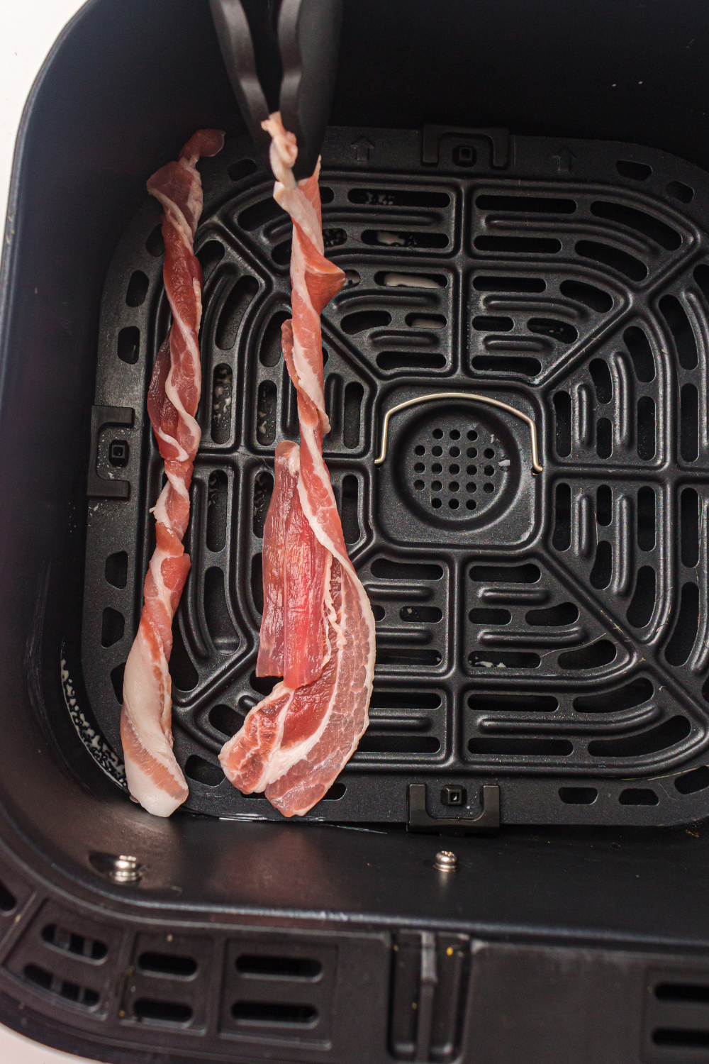 Hands twisting bacon strip into a tight spiral rope shape with opposite hand rotation to create a firm twisted bacon design that holds its shape during air frying and results in crispy caramelized texture