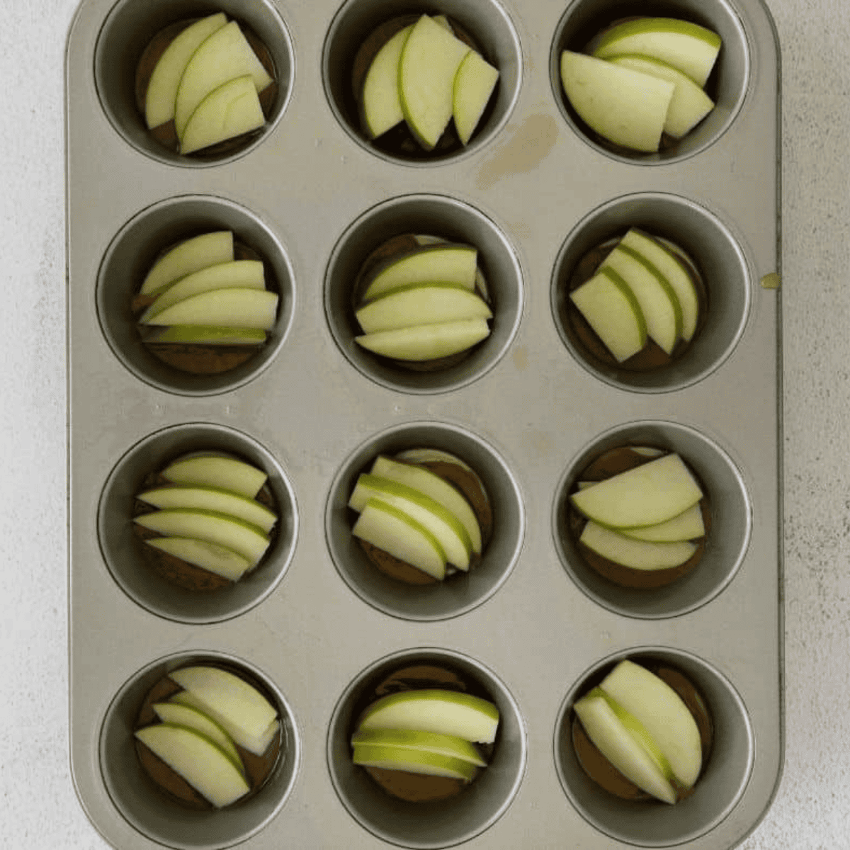 Placing thin Granny Smith apple slices into muffin tins over a brown sugar butter mixture, gently pressing them in to form the base layer for Amish Apple Cake.