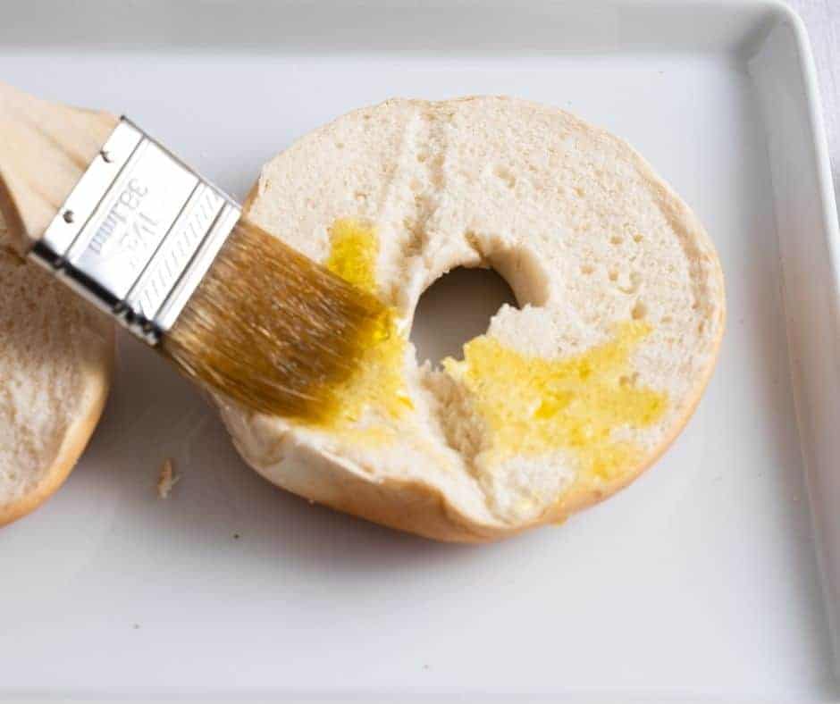 Halved bagels on a flat surface being brushed with olive oil.