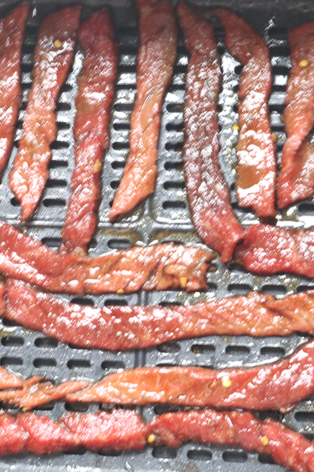 Marinated beef strips arranged in a single layer in an air fryer basket, not overlapping, ready to dehydrate into jerky