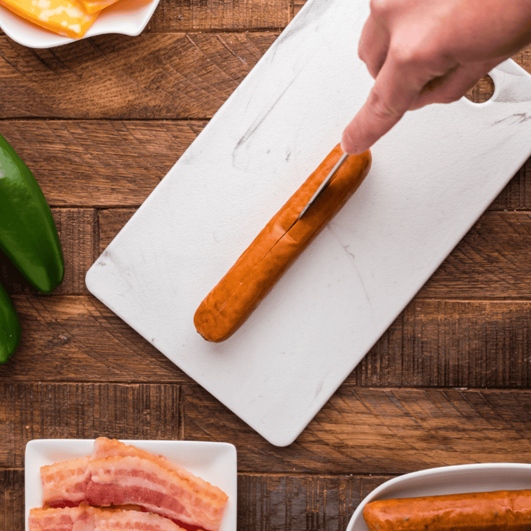 Hot dog sliced lengthwise and stuffed with cheddar cheese and a jalapeno slice on a cutting board