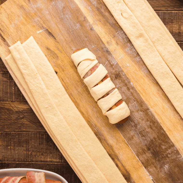 Wrap with dough step: Crescent dough strip wound around a bacon-wrapped hot dog to form a pretzel dog