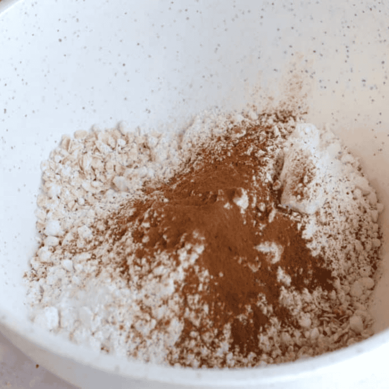 Melted butter in bowl next to a large bowl of flour, oats, cinnamon, baking soda, and salt whisked together for cookie dough preparation.