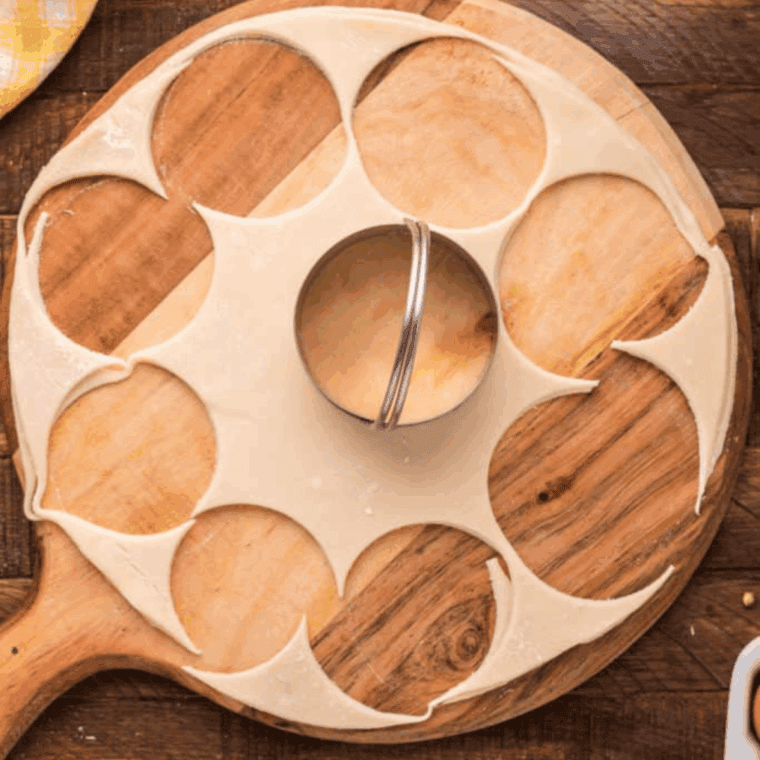 Stacked pie crusts on a wooden cutting board with circles being cut using a mason jar lid for meat pies.