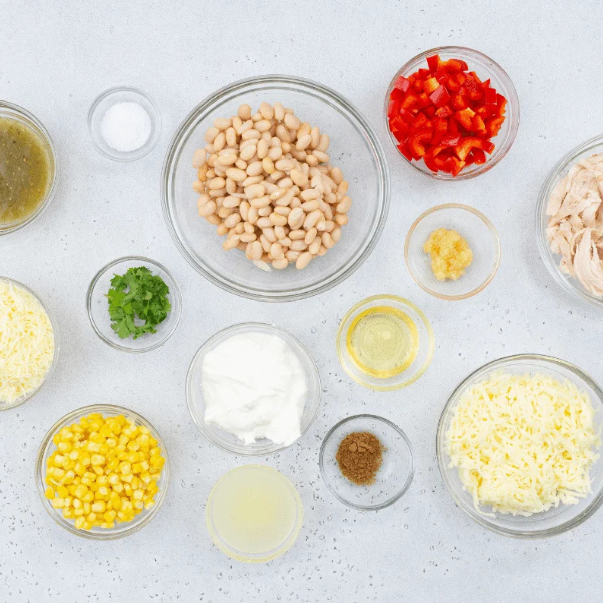 White beans, sour cream, shredded rotisserie chicken, cheddar, mozzarella, salsa verde, corn, red bell pepper, garlic, lime, and cumin arranged on a kitchen counter