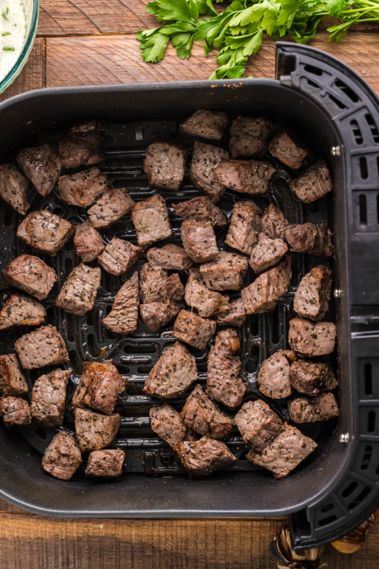 Seasoned steak bites arranged in a single layer in an air fryer basket, spaced apart for even cooking and browning.