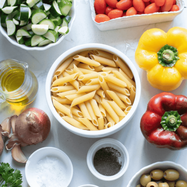 Flat lay of pasta salad ingredients: dry pasta, cherry tomatoes, zucchini, red and yellow bell peppers, red onion, garlic, olives, lemon, parsley, olive oil.