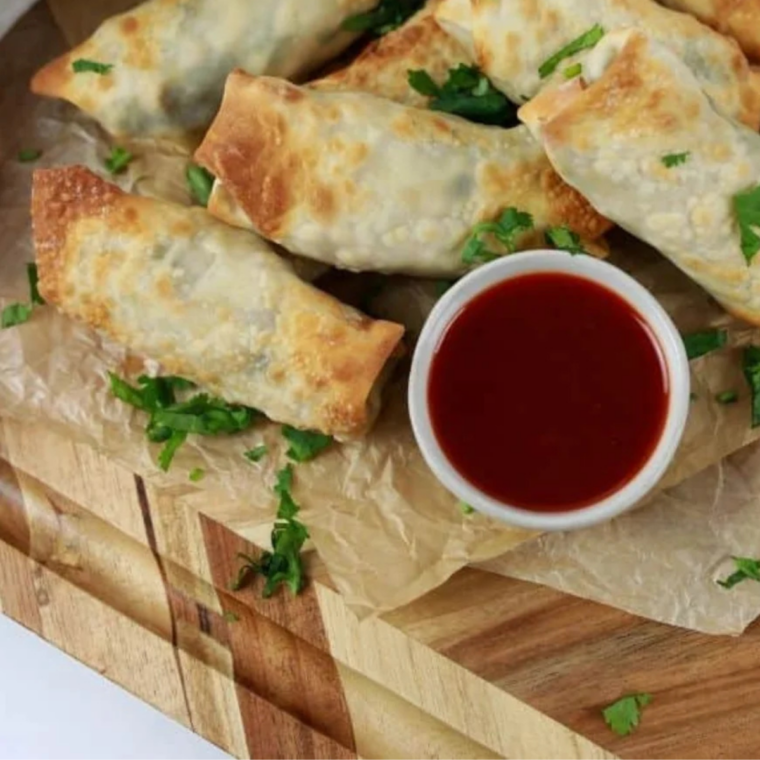 Air fryer chicken cilantro egg rolls stacked on a white plate with a bowl of sweet and sour sauce