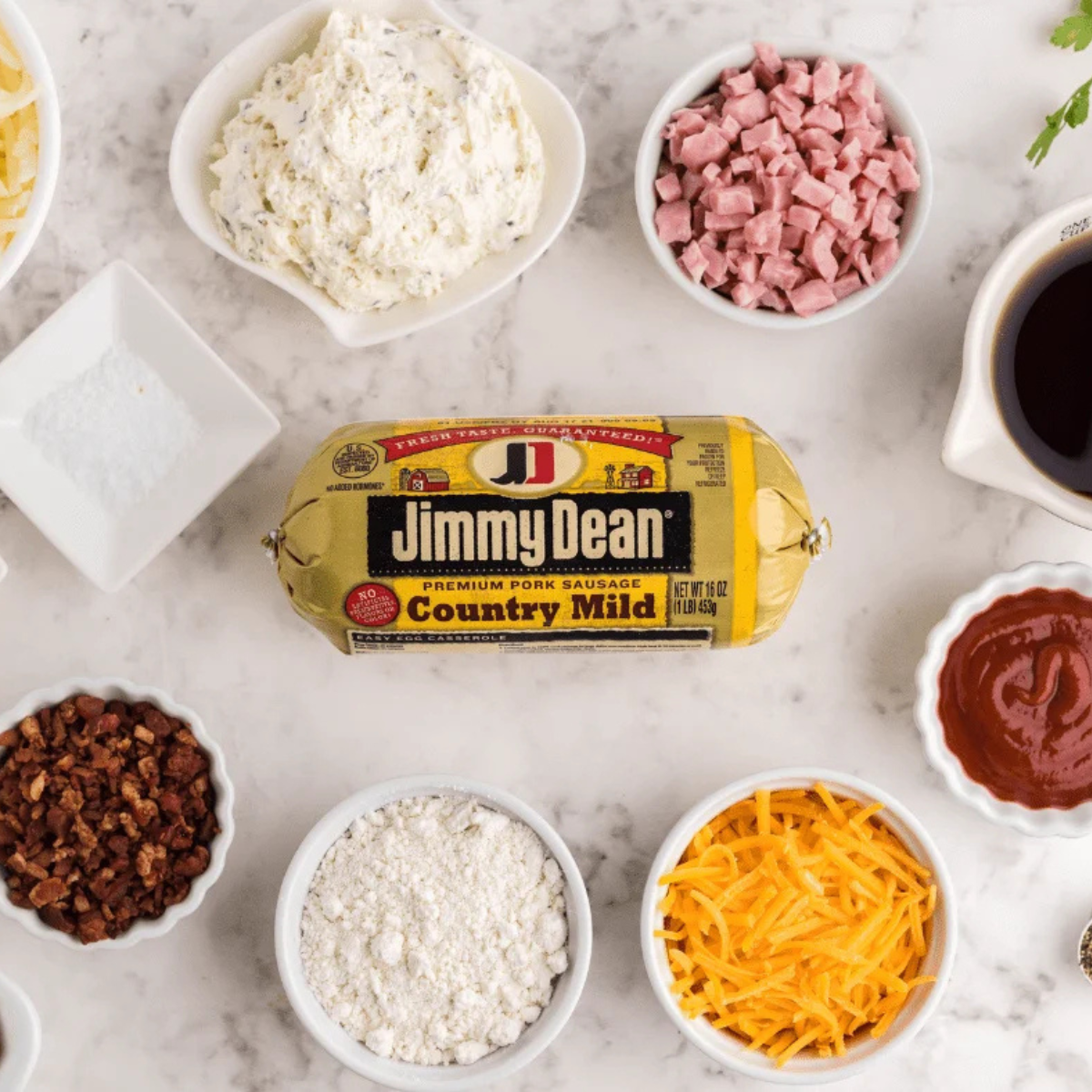 Ingredients for Air Fryer Sausage Balls arranged on a counter: ground pork sausage, shredded hash browns, Bisquick, bacon, diced ham, cheddar cheese, eggs, and cream cheese