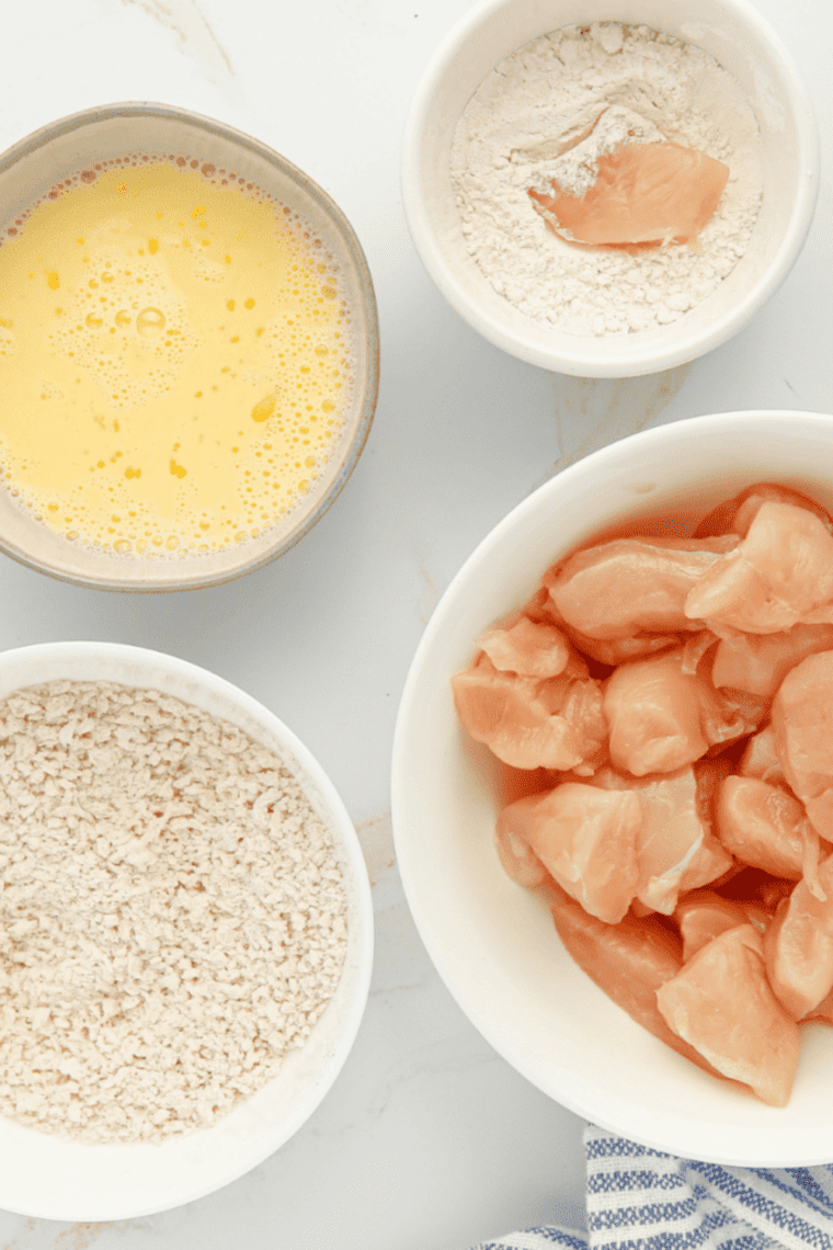 Air fryer basket sprayed with cooking spray alongside bowls of flour, egg and milk mixture, and seasoned breadcrumb coating ready for popcorn chicken.