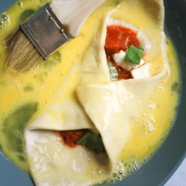 Folded pizza cannoli being brushed with egg wash in a shallow bowl.