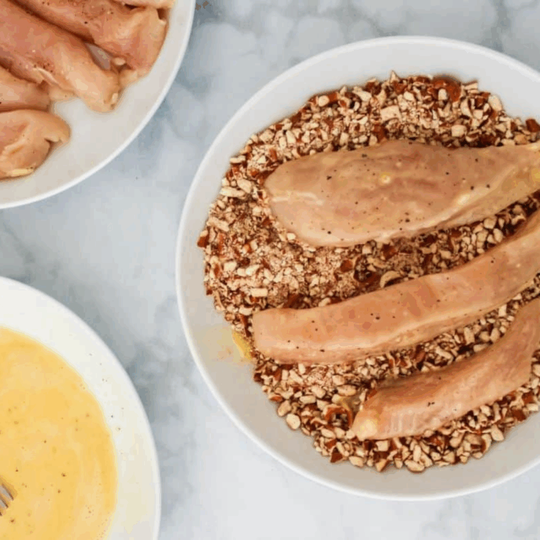 Seasoned chicken tenders with salt and pepper, flour in one dish, beaten eggs with Dijon mustard in another, and crushed pretzels in a third dish ready for breading.