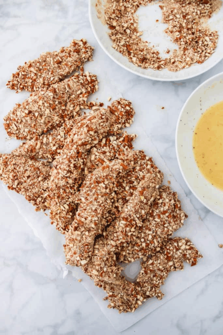 Chicken tender being coated step-by-step: first in flour, then egg and mustard mixture, and finally covered with crushed pretzels.