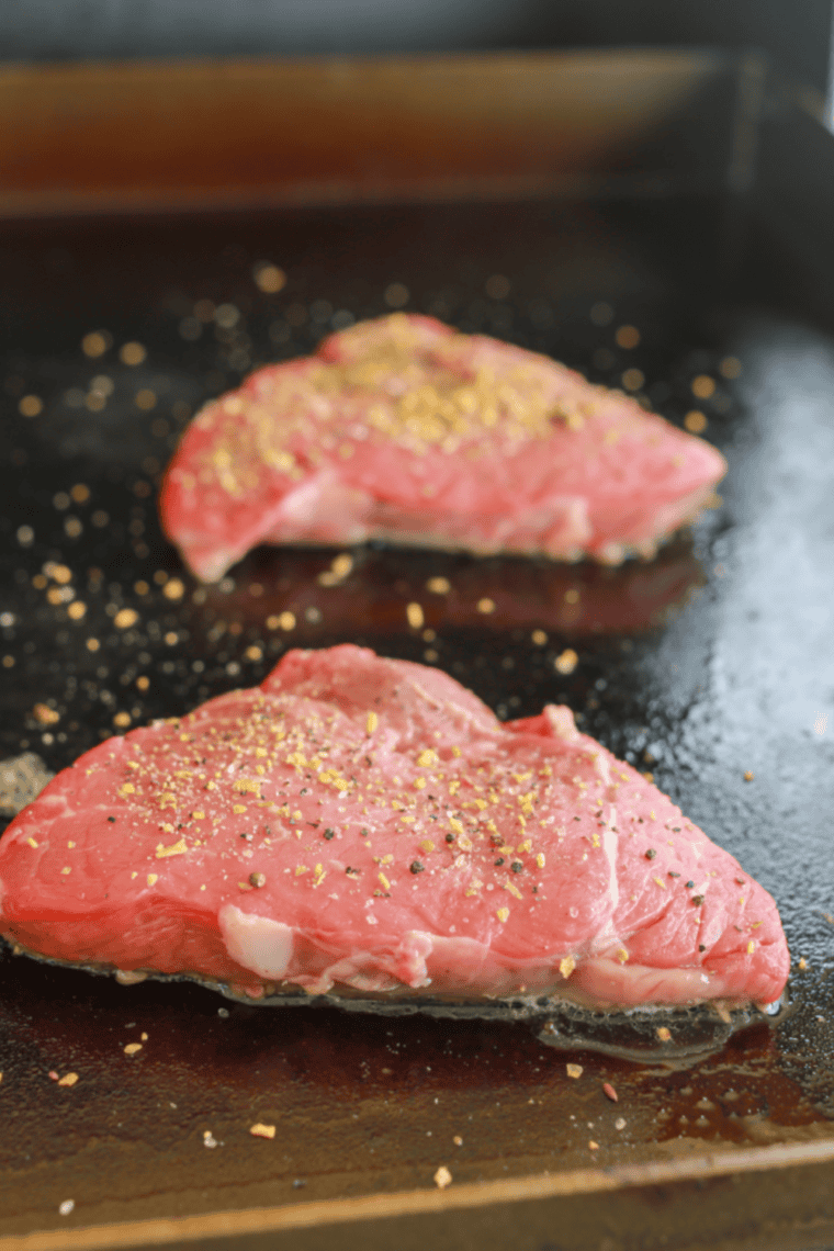 Two sirloin steaks searing on a hot Blackstone griddle with visible steam and a dark brown crust forming.