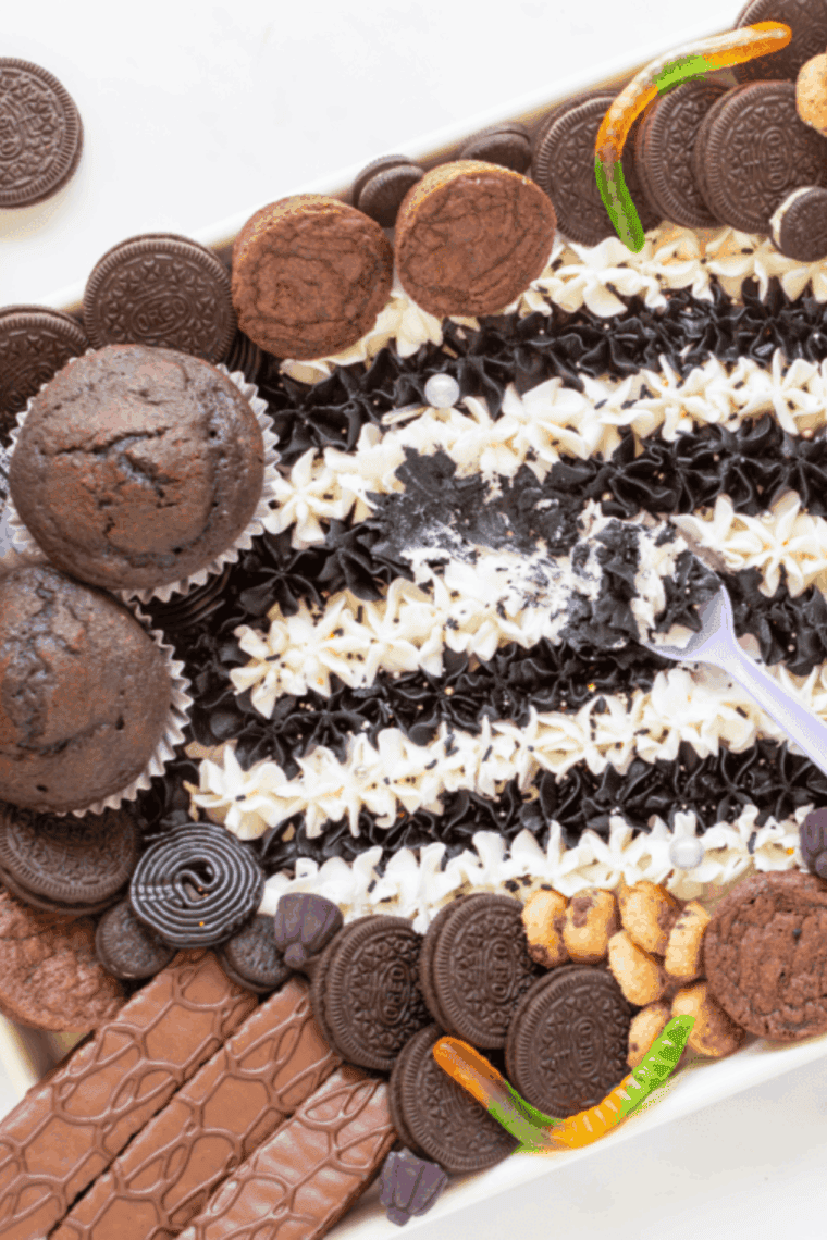 Beetlejuice-inspired buttercream board with black, white, purple, and green frosting, decorated with cupcakes, brownies, and candy.