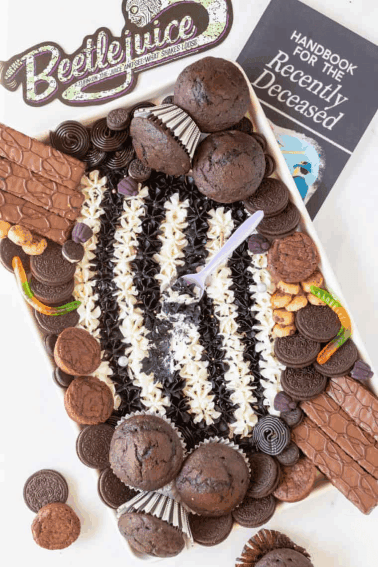 Beetlejuice-inspired buttercream board with black, white, purple, and green frosting, decorated with cupcakes, brownies, and assorted candy.
