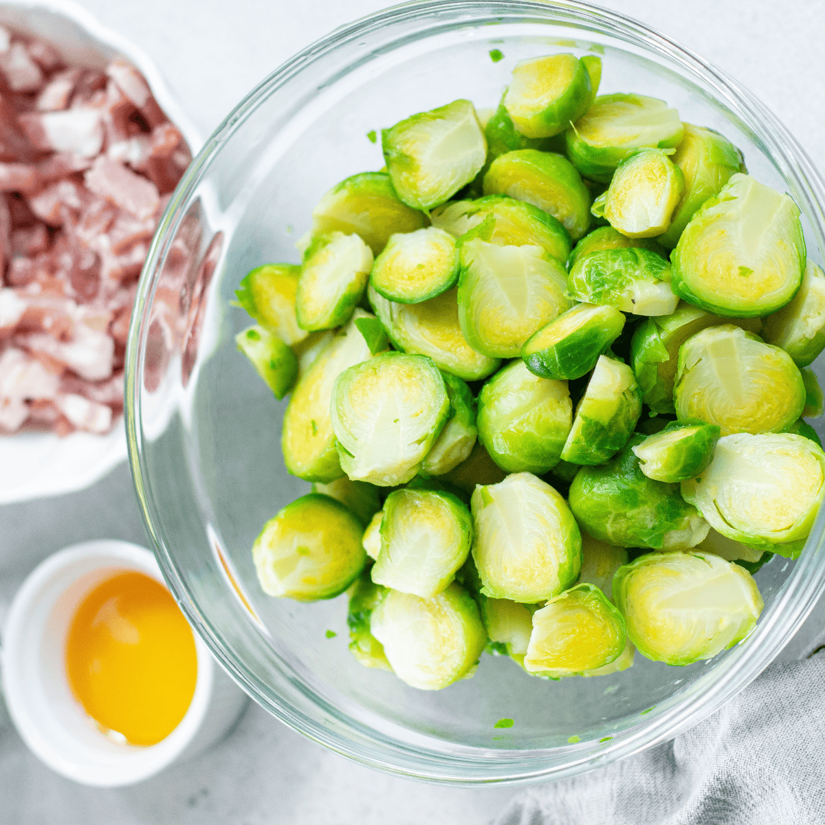 Fresh Brussels sprouts trimmed, halved, and ready for cooking, with chopped bacon pieces on the side.