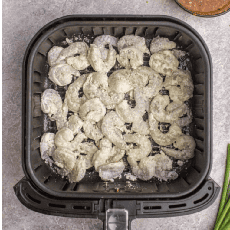 Coated shrimp in a single layer in the air fryer basket sprayed with oil