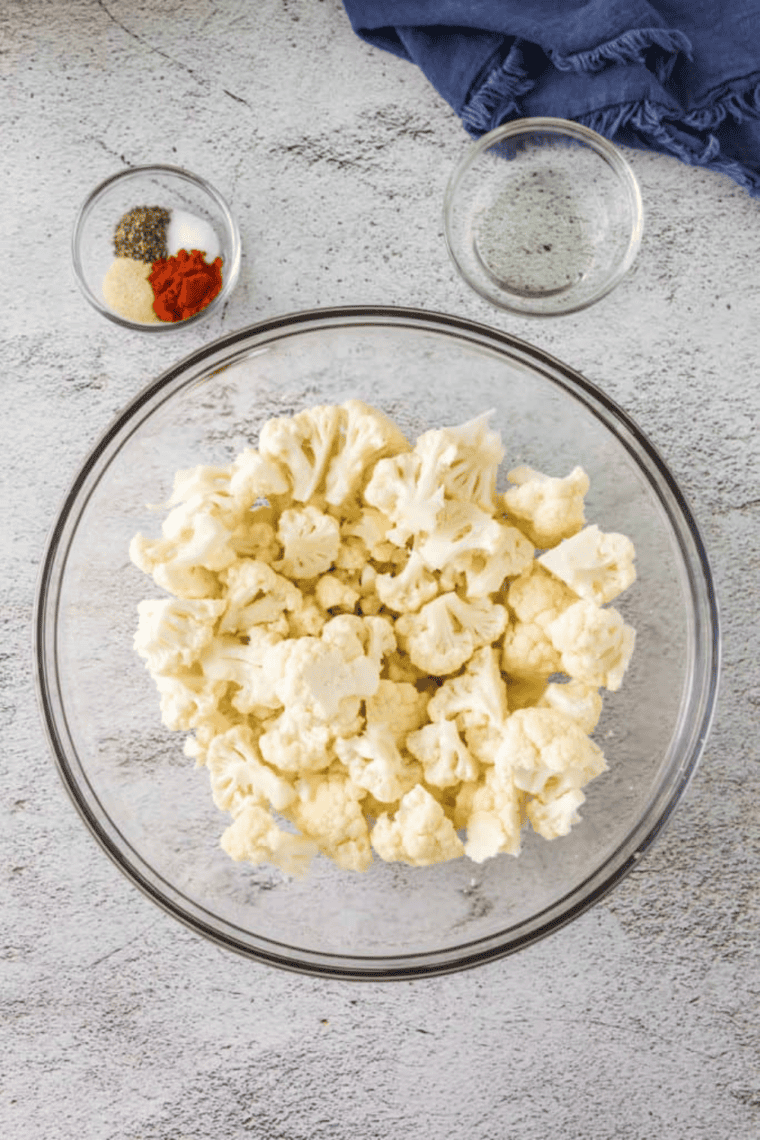 Cauliflower florets in a large bowl being tossed with minced garlic, olive oil, kosher salt, and freshly ground black pepper for even coating before roasting.