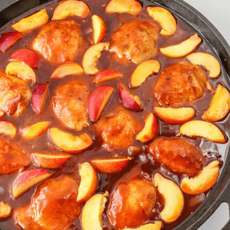 Jar of golden whiskey peach BBQ sauce with fresh peaches and spices on a wooden board.