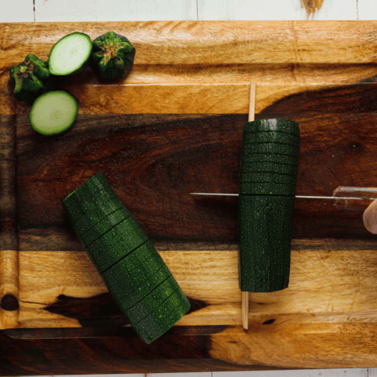 Medium zucchini being sliced into thin, parallel cuts, fanning slightly but staying intact at the base.