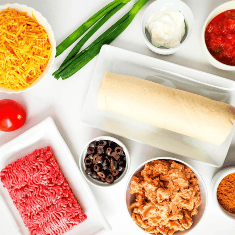 Taco pizza ingredients laid out: refrigerated pizza dough, ground beef, taco seasoning, refried beans, salsa, cheddar cheese, olives, tomato, and green onions