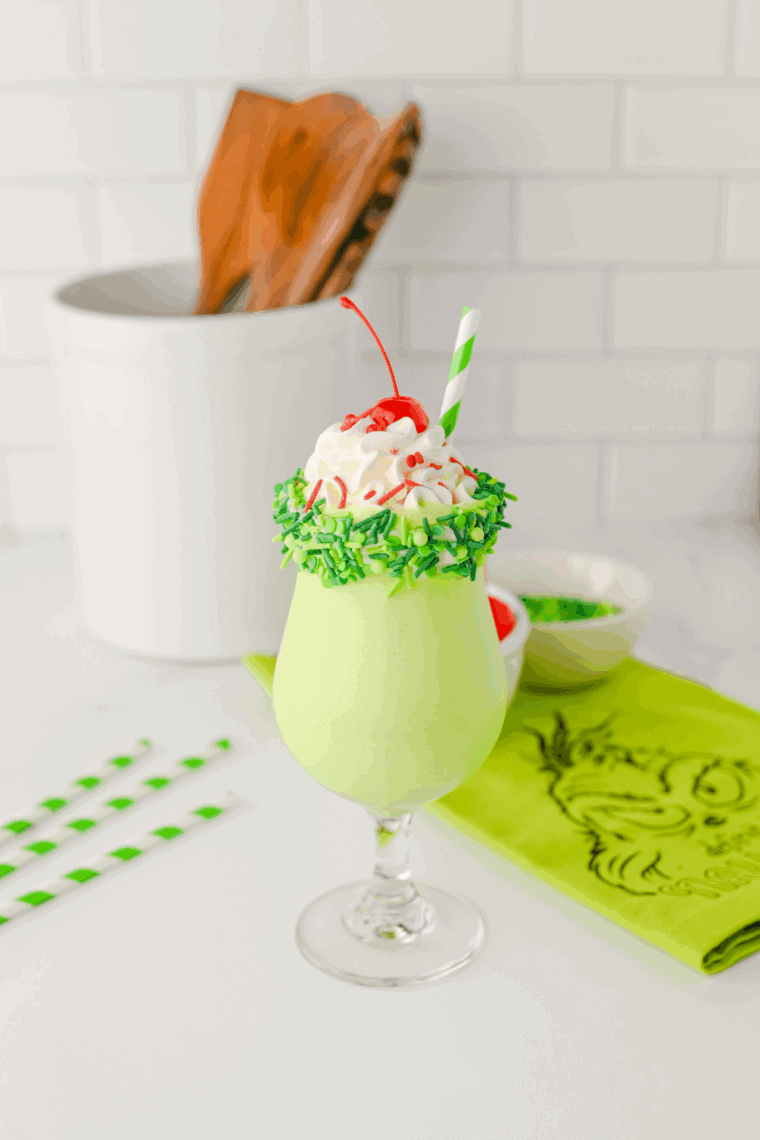 Green Grinch milkshake in a glass, topped with whipped cream and colorful holiday sprinkles.