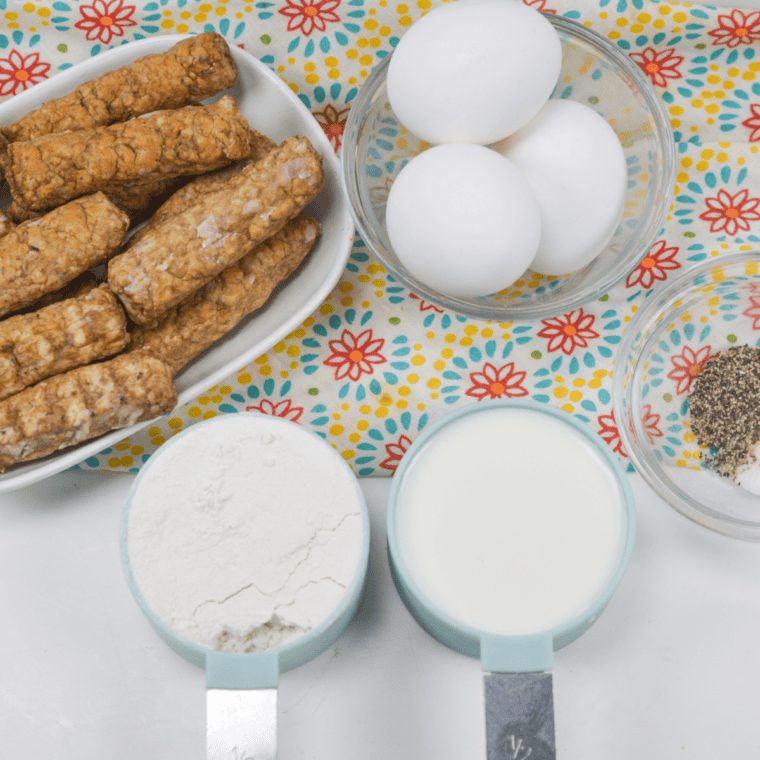 Ingredients for air fryer toad in the hole laid out on a wooden board: pork sausages, flour, eggs, milk, and fresh herbs