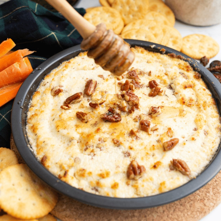 Baked goat cheese dip with honey, pecans, and fresh thyme in a white baking dish surrounded by toasted French bread slices