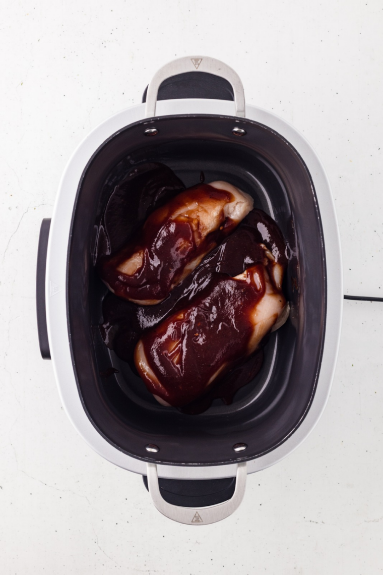Barbecue sauce mixture being poured over seasoned chicken breasts in the slow cooker