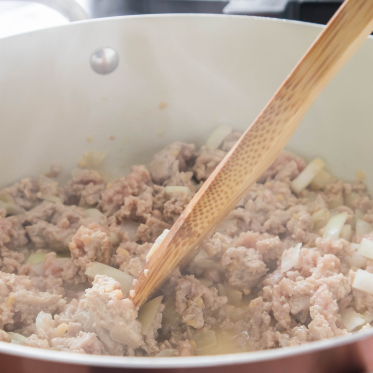 Raw ground beef and beef broth in a saucepan being broken up with a potato masher