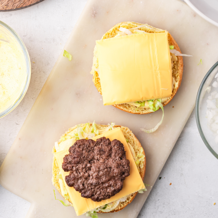  Assembling a homemade Big Mac with layers of special sauce, shredded lettuce, American cheese, beef patties, pickles, and onions between a three-part sesame seed bun.