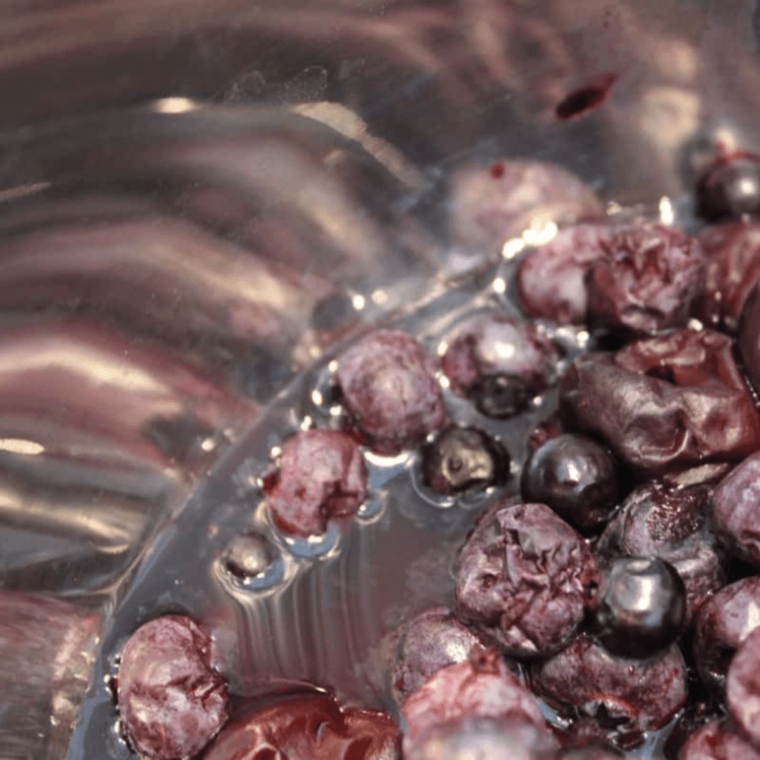 Thawed blueberries in a strainer being rinsed under running water, with stems and debris being removed.