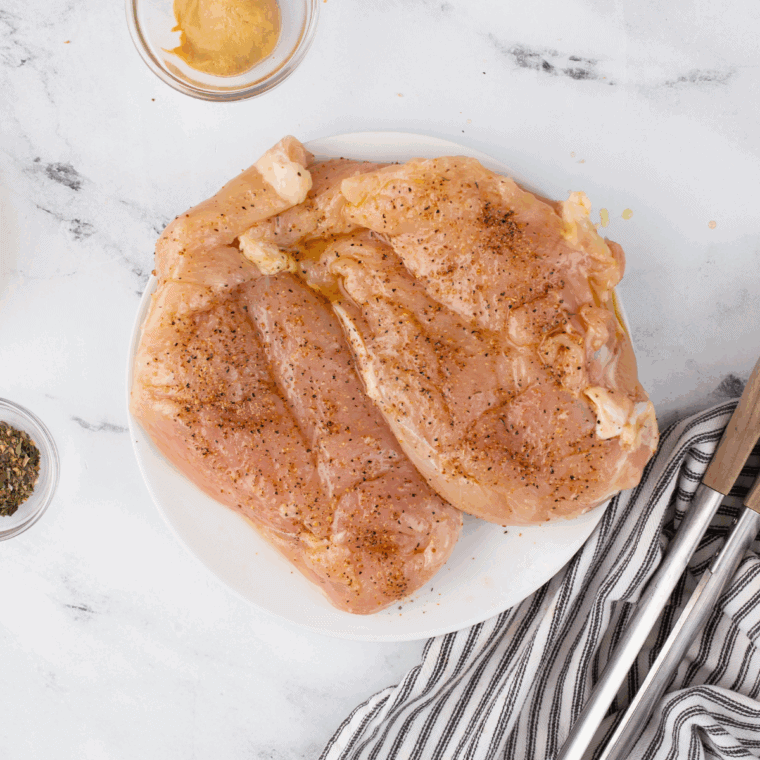 Raw chicken breasts brushed with olive oil and seasoned with salt and pepper on a cutting board.