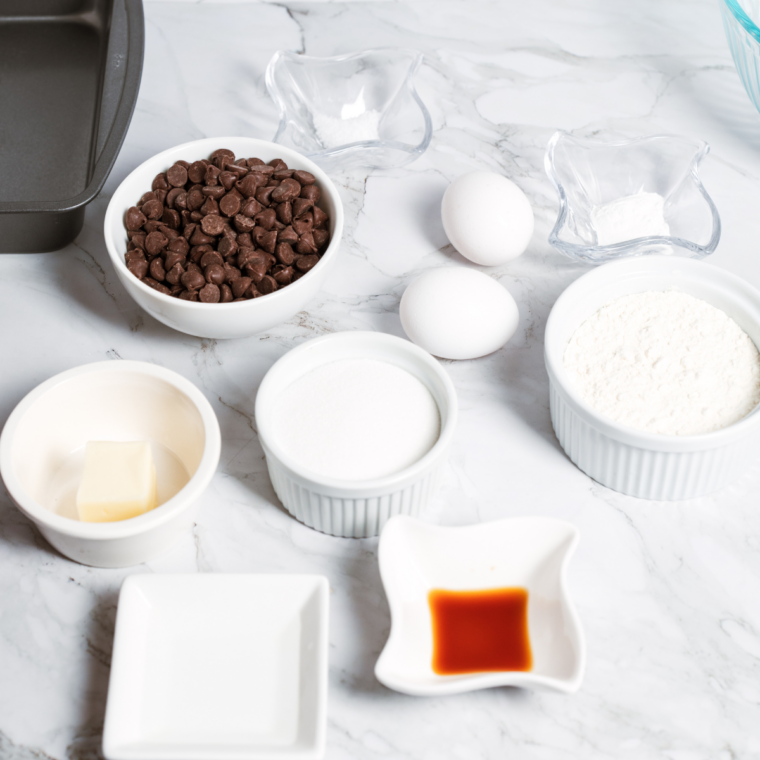 Overhead flat lay of brownie ingredients including cocoa powder, eggs, butter, and chocolate chunks